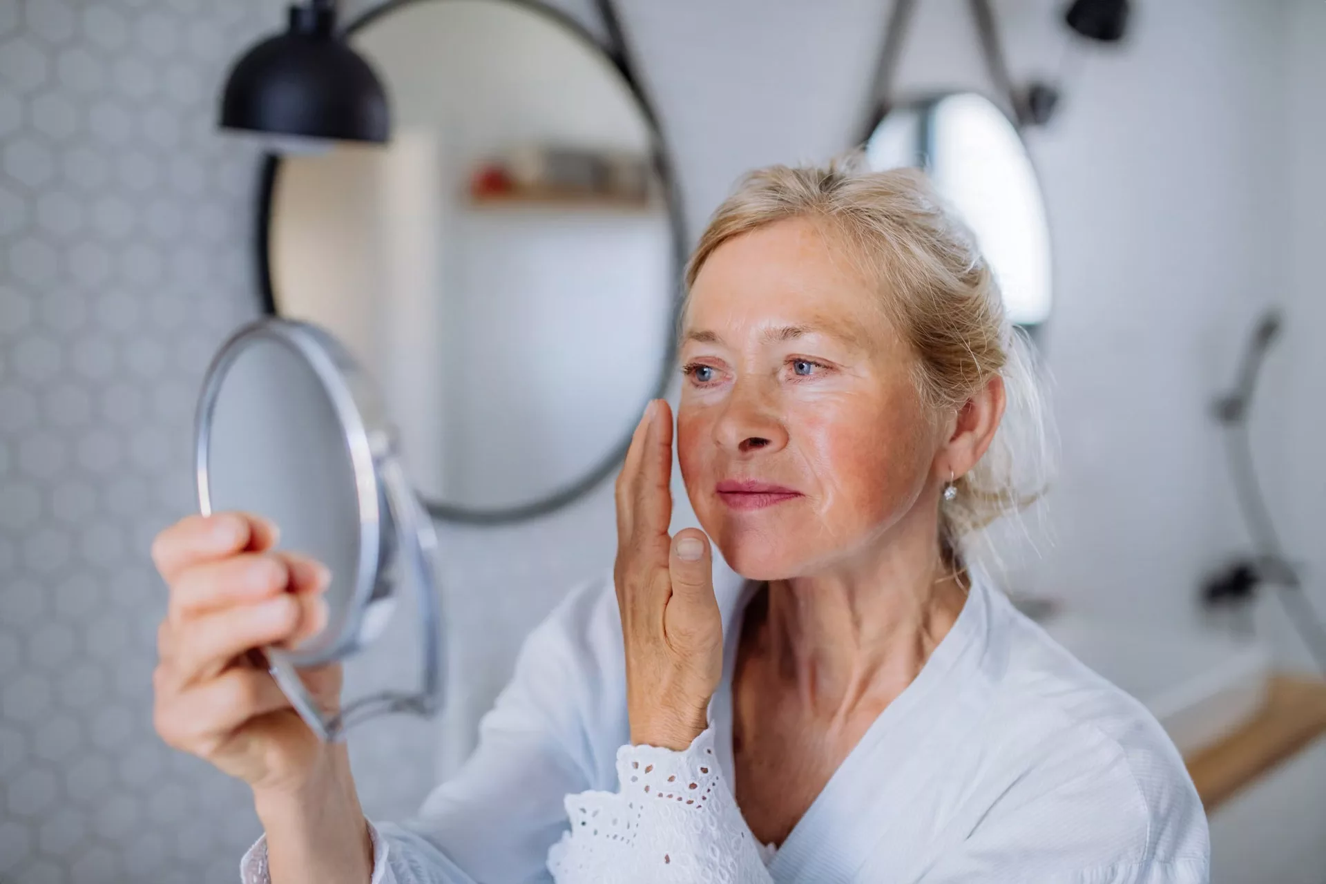 une femme agee en peignoir regardant le miroir et appliquant une creme pour le visage dans la salle de bain concept de soins de la peau